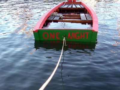 Ein grün-rotes Ruderboot, an einem Seil befestigt, ist im Begriff, unterzugehen. Es ist viel Wasser im Boot. Auf dem Heck steht 'one night'.