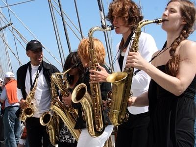 Fünf Saxophon-Spieler stehen nebeneinander auf einem Segelschiff und spielen Saxophon. Im Vordergrund spielt eine Frau im schwarzen, ärmellosen Kleid.