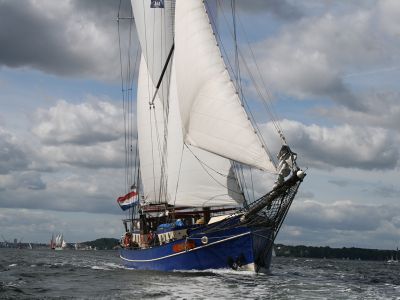 Ein Segelschiff mit blauem Rumpf und vollen weißen Segeln segelt auf dem Wasser. Am Himmel sind weiß-graue Wolken.