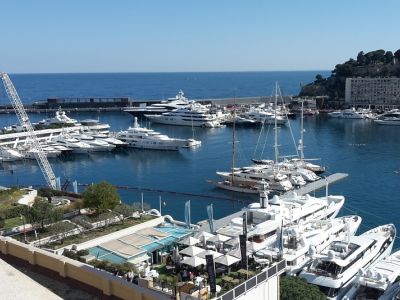 Im Hafen von Monaco liegen weiße Segelboote. Im Hintergrund das blaue Meer und Felsen, die die Bucht umschließen.