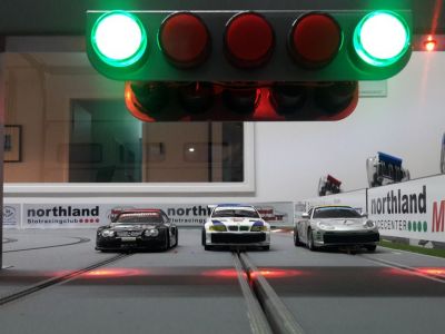 Drei Miniatur-Rennautos stehen nebeneinander am Start einer Slotcar-Bahn. Über den Autos leuchten zwei grüne Startampeln.