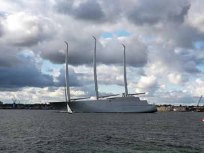 Eine graue Mega-Segelyacht mit drei Masten ohne Segel schwimmt vor der Küste.