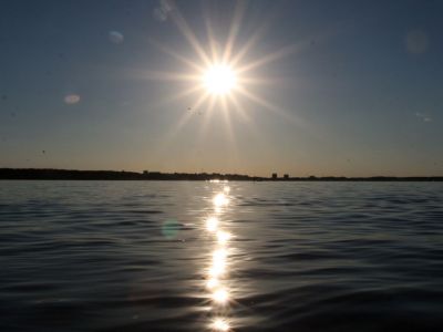 Die Sonne steht tief über der Kieler Förde. Sie spiegelt sich auf der Wasseroberfläche. Am Horizont ist Land zu sehen.