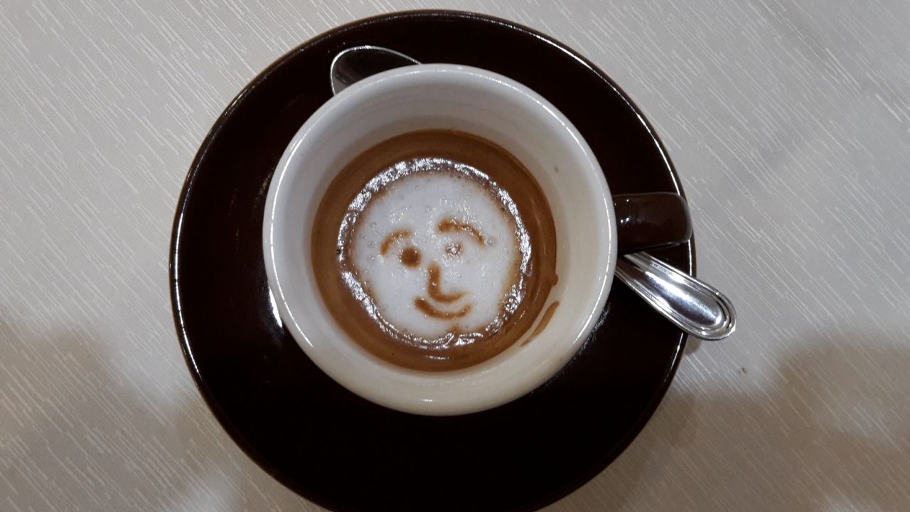 In einer schwarzen Kaffeetasse sieht man Milchschaum. Darauf ist mit Kakaopulver ein Smiley getreut