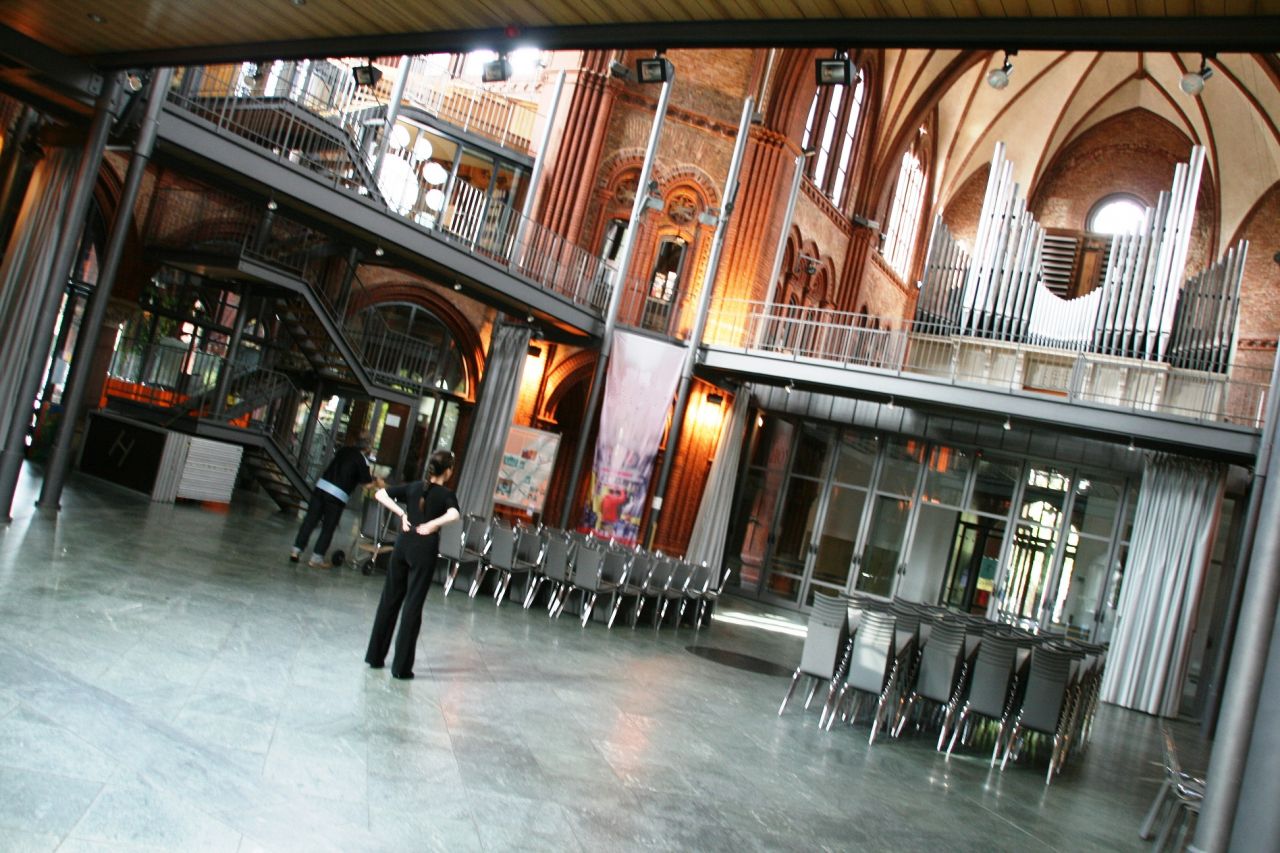 Im leeren Raum in einer Kirche steht eine Person uns schaut zur Orgel hinauf. Zwei Dinnertische mit Stühlen stehen im Raum.