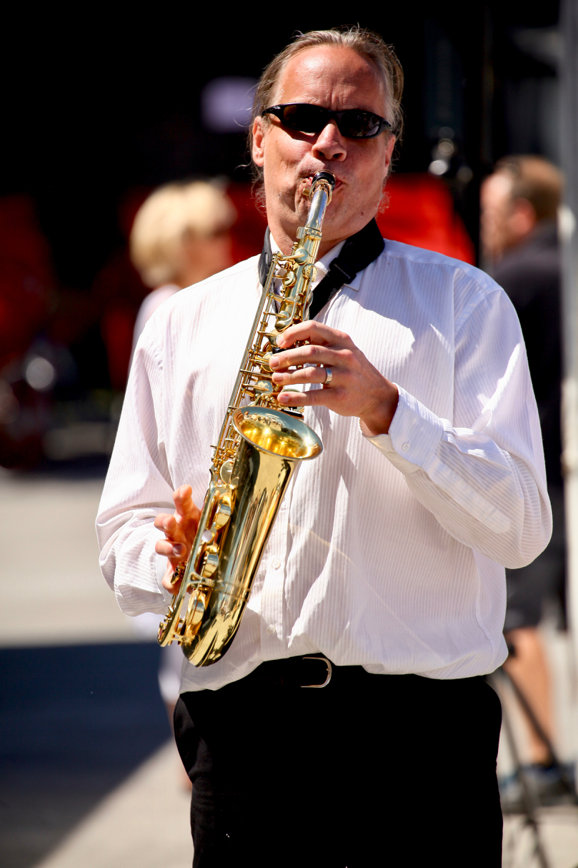 Ein Mann spielt Saxophon. Er trägt eine dunkle Sonnenbrille, ein weißes Hemd und eine schwarze Hose.