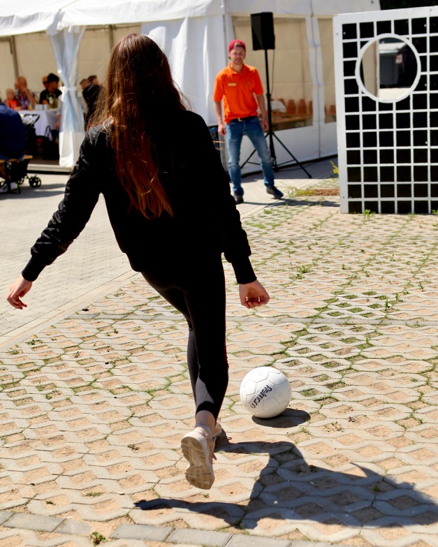 Im Hintergrund ist eine Torwand zu sehen, daneben ein Mann in orange farbenem Poloshirt. Eine junge Frau mit offenen dunklen, langen Haaren nimmt Anlauf, um einen weißen Lederball auf die Torwand zu schießen.