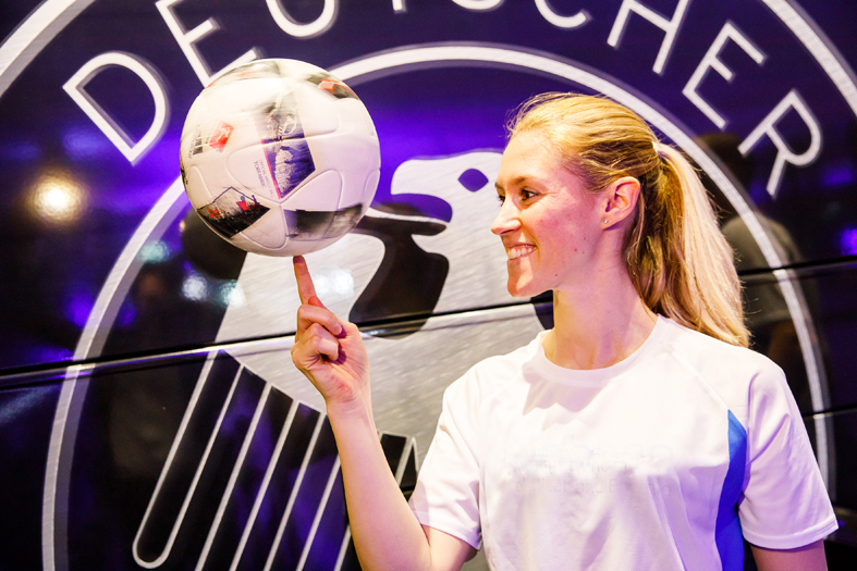 Eine Frau mit blonem Zopf in weißem Trikot lässt einen Lederfußball auf dem Zeigefinger rotieren. Im Hintergrund ist teilweise der Adler aus dem Wappen des Deutschen Fußballbundes zu sehen.